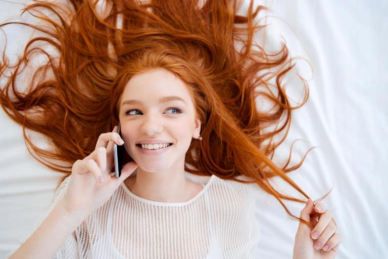 Ung smilende rødhåret pige der ligger på seng og taler i mobil
