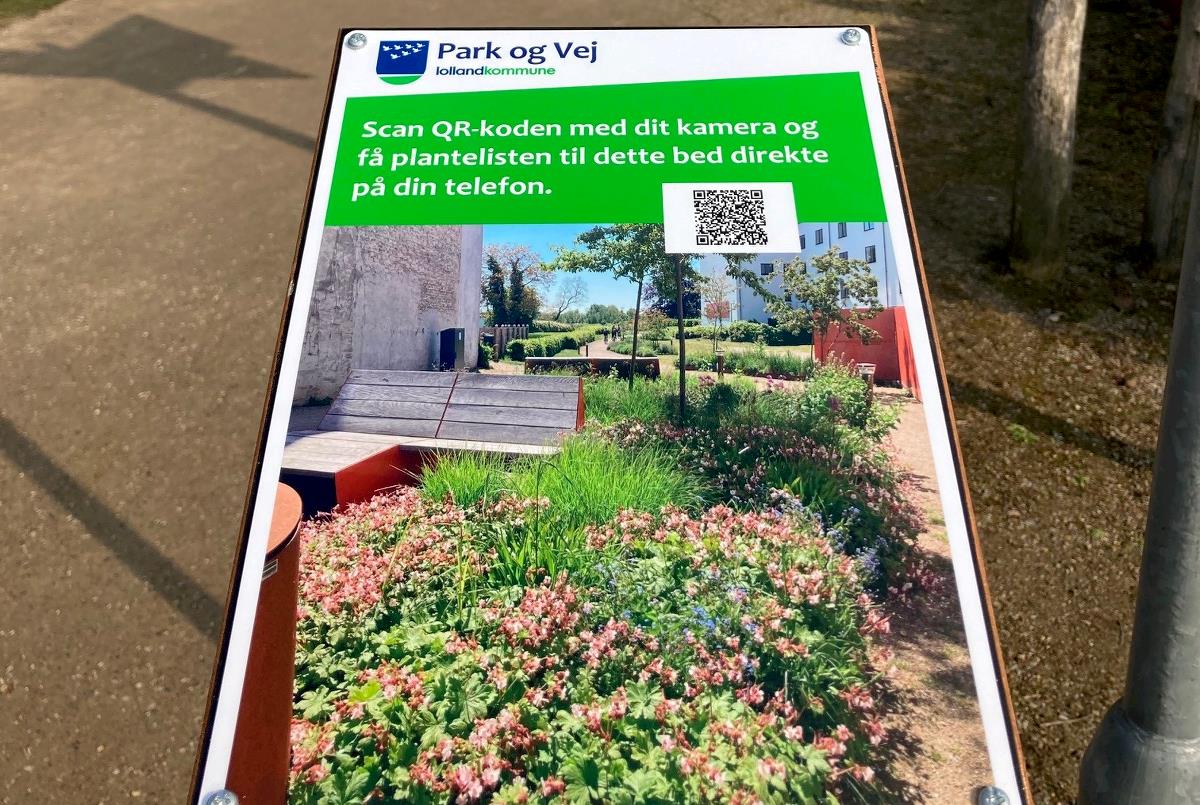 et nær billede af et skilt, hvor der er et foto af et blomsterbed samt en QR kode.